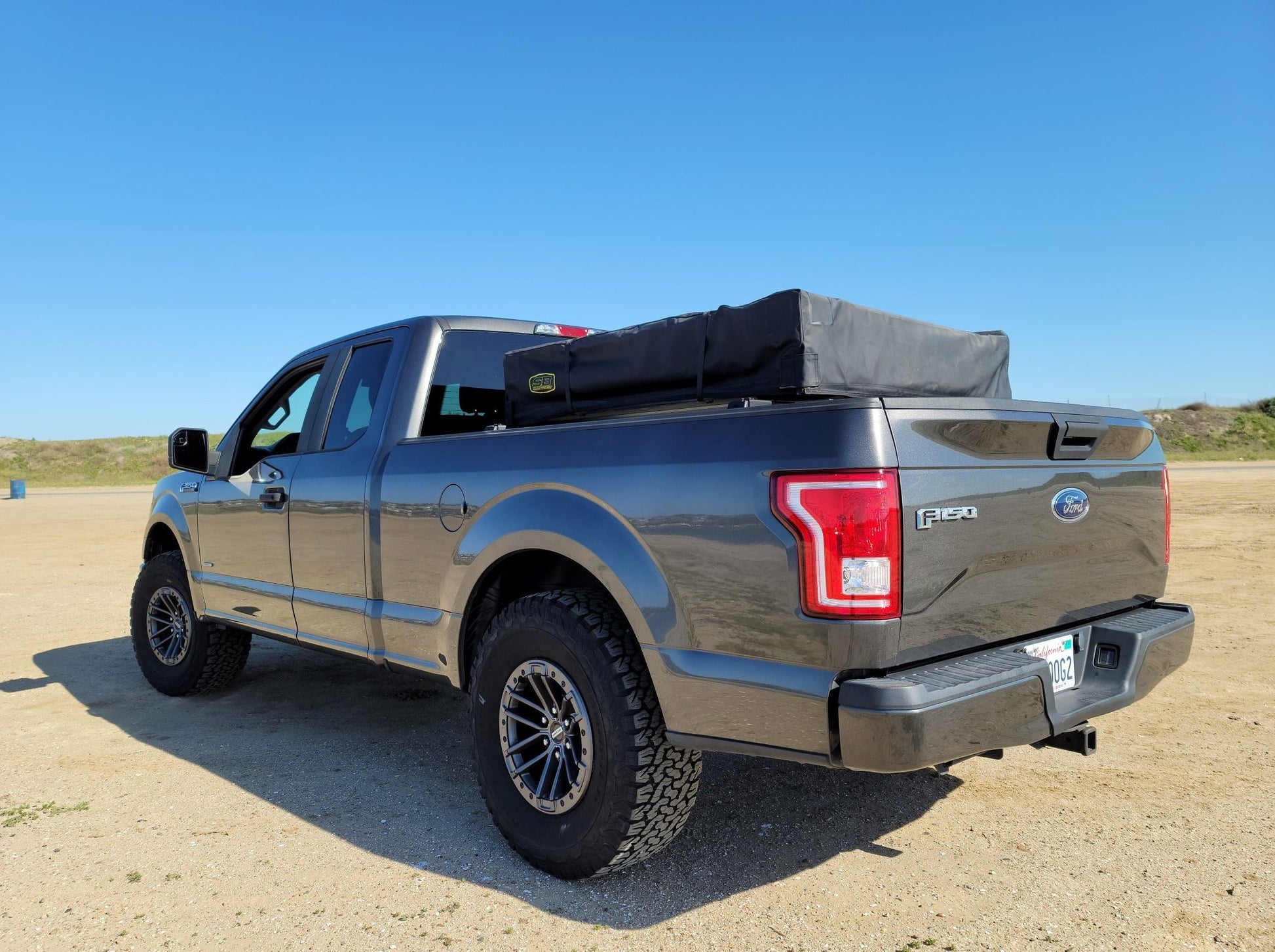 2015+ F-150 Low Profile Rooftop Tent Rack Kit by Range Industries, featuring the rear view of a gray F-150 parked on the beach with a rooftop tent in the truck bed.