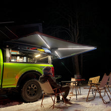 Load image into Gallery viewer, Nighttime camping scene featuring a Dirtbox Overland Free Standing 270-Degree Awning with integrated LED lighting, illuminating the area around a camper enjoying the outdoors.