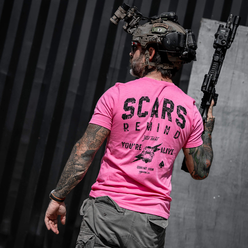Tattooed man in a pink t-shirt saying 'Scars Remind You That You're Alive' by Combat Iron Apparel in an urban setting.