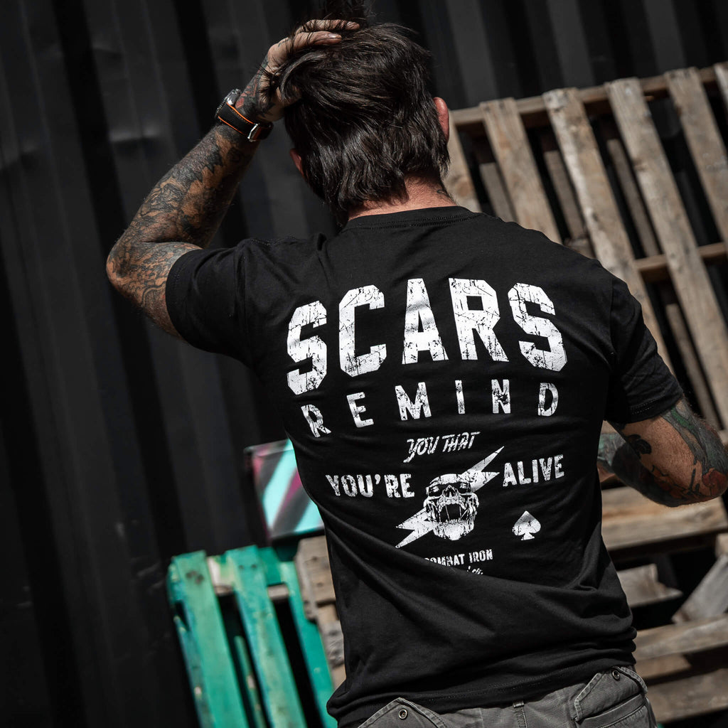 Close-up of a tattooed man wearing a black t-shirt with 'Scars Remind You That You're Alive' by Combat Iron Apparel.