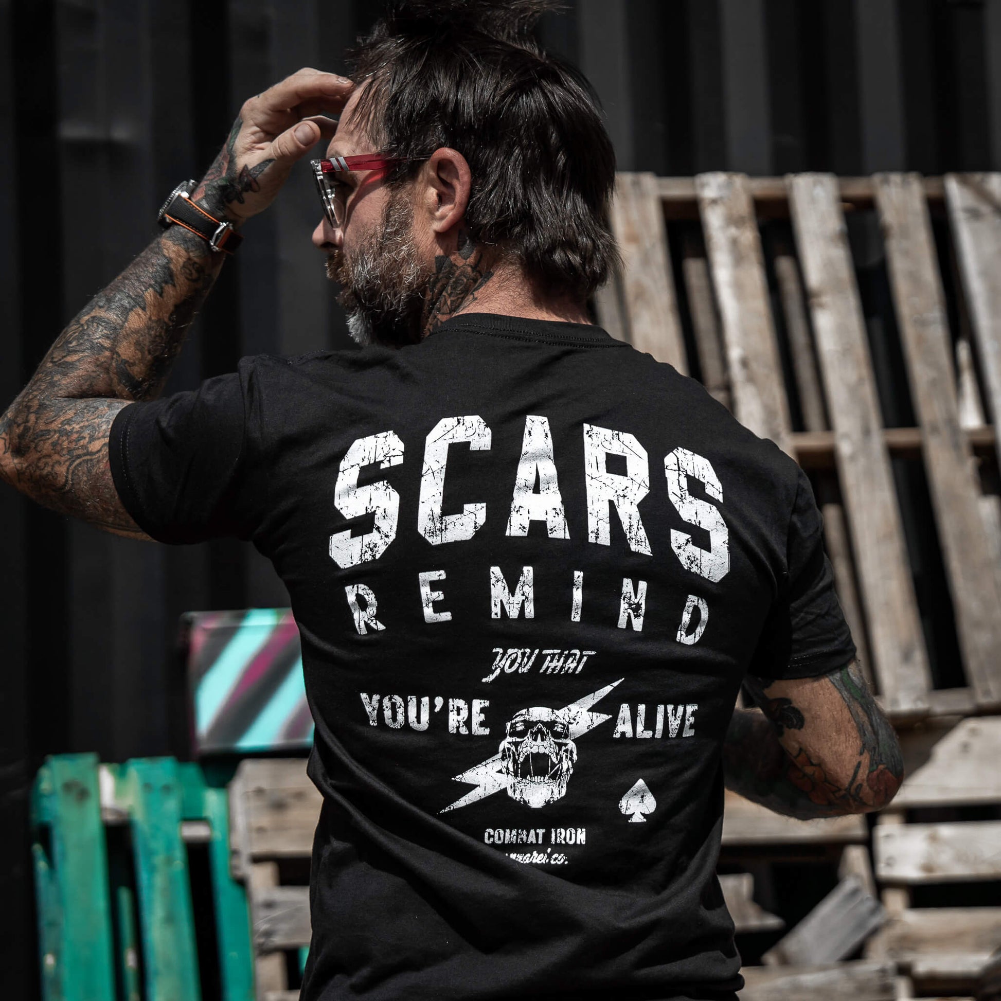 Man wearing a black t-shirt with the phrase 'Scars Remind You That You're Alive' by Combat Iron Apparel, showing a tattooed arm.