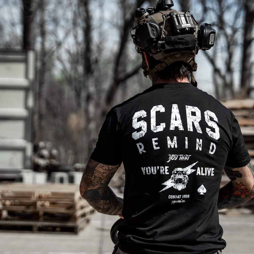 Tattooed man in a tactical setting wearing a black t-shirt with 'Scars Remind You That You're Alive' by Combat Iron Apparel.