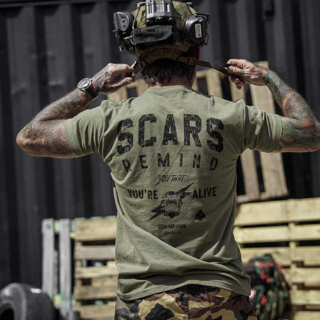 Close-up of a tattooed man holding gear while in an olive green t-shirt with 'Scars Remind You That You're Alive' by Combat Iron Apparel.