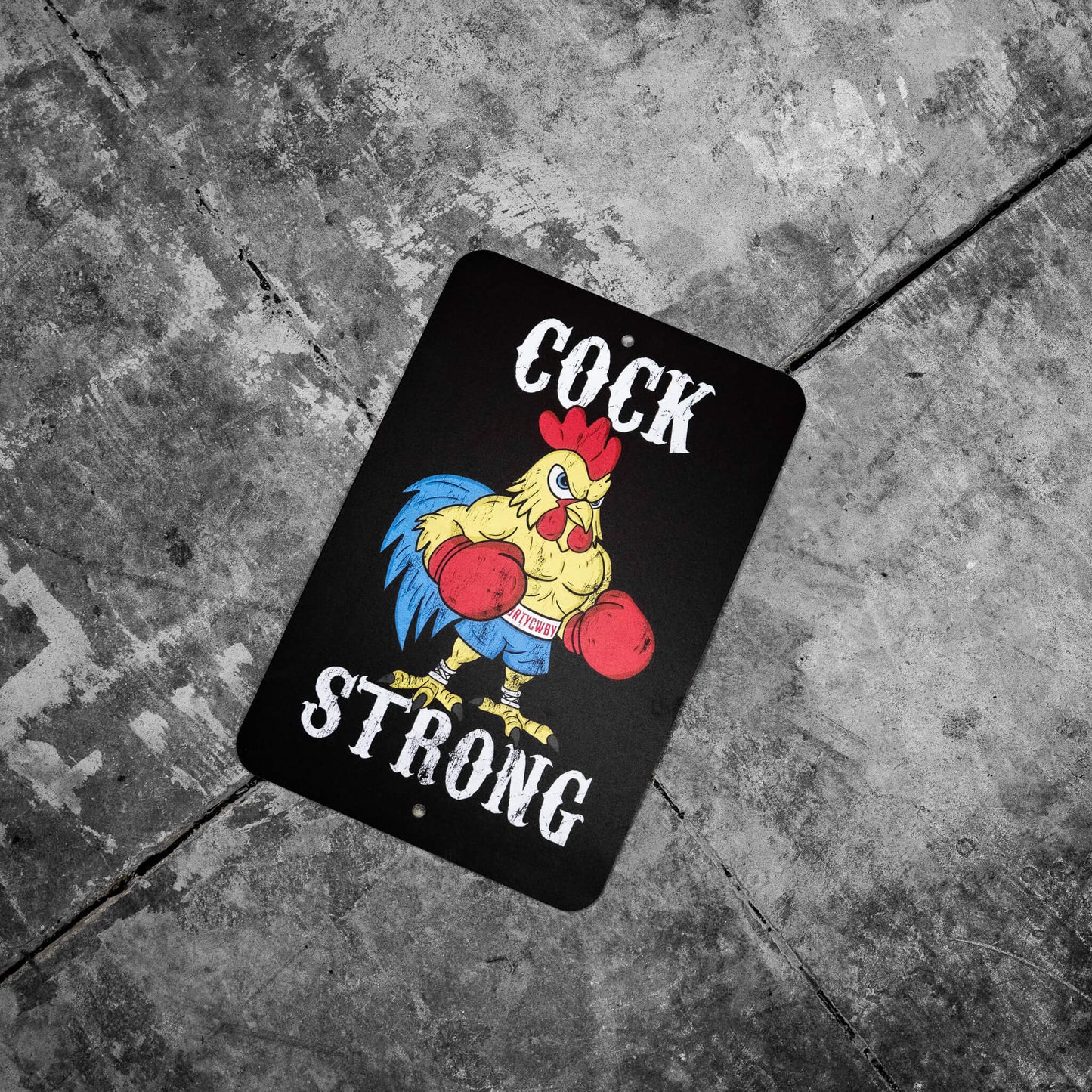 Cock Strong metal sign by Combat Iron Apparel resting on a rugged stone floor, featuring a bold rooster in red boxing gloves.