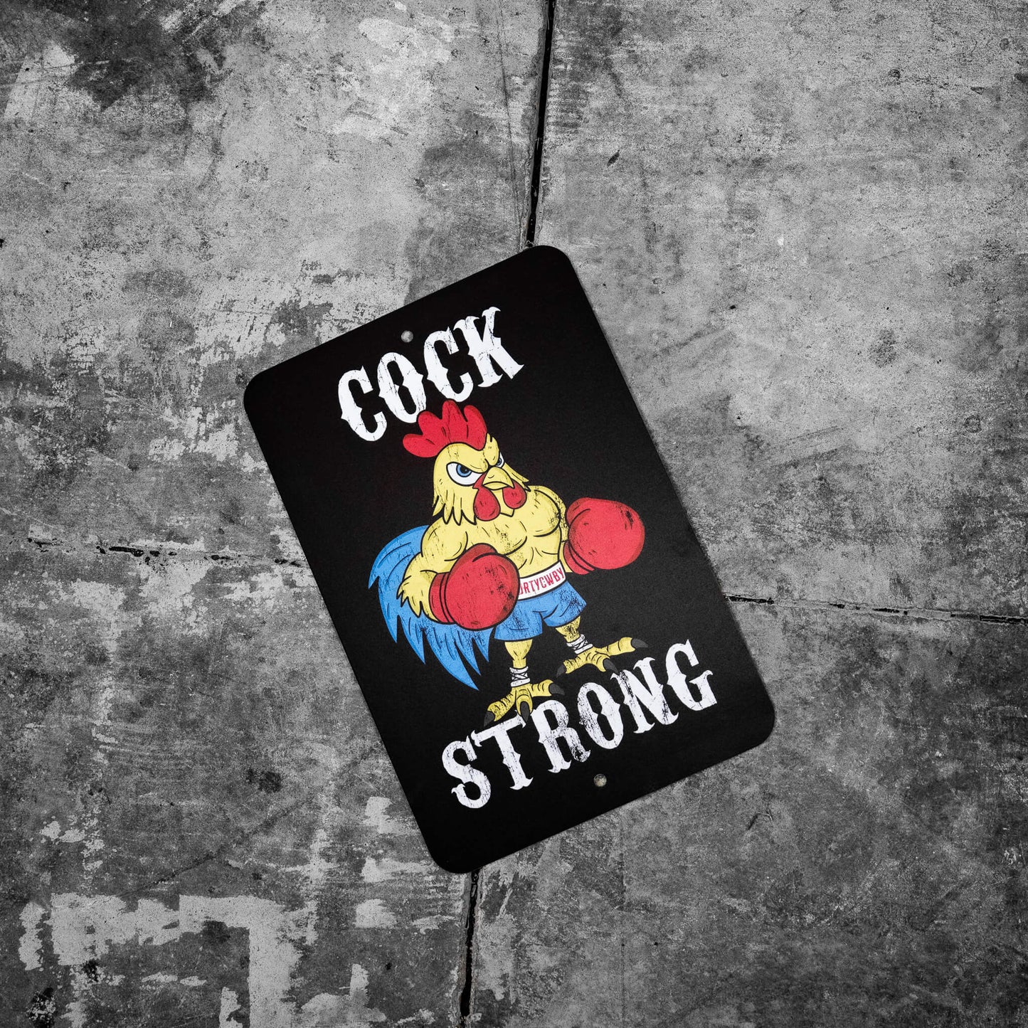 Cock Strong metal sign by Combat Iron Apparel laid on a gritty stone surface, highlighting its vibrant rooster design.