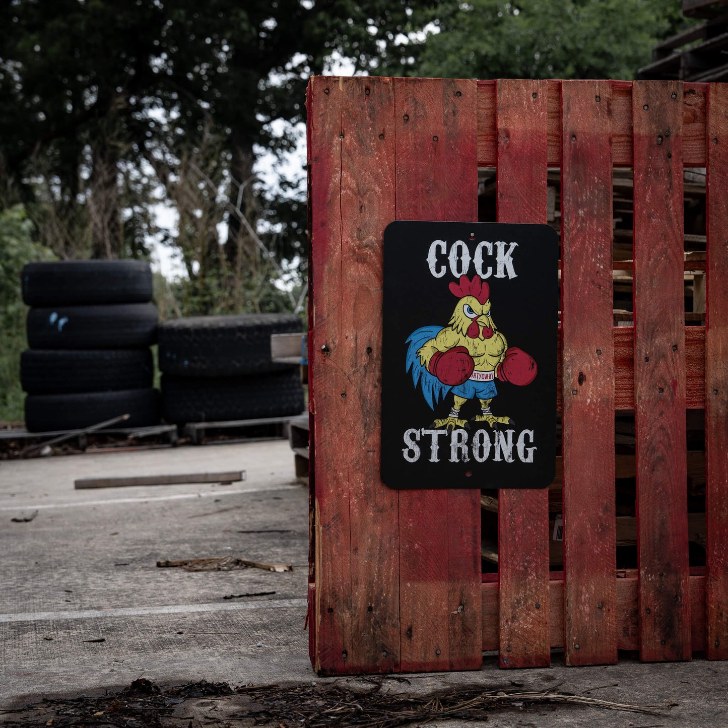 Cock Strong wall-mounted sign by Combat Iron Apparel decoratively placed in an outdoor setting with a wooden fence.