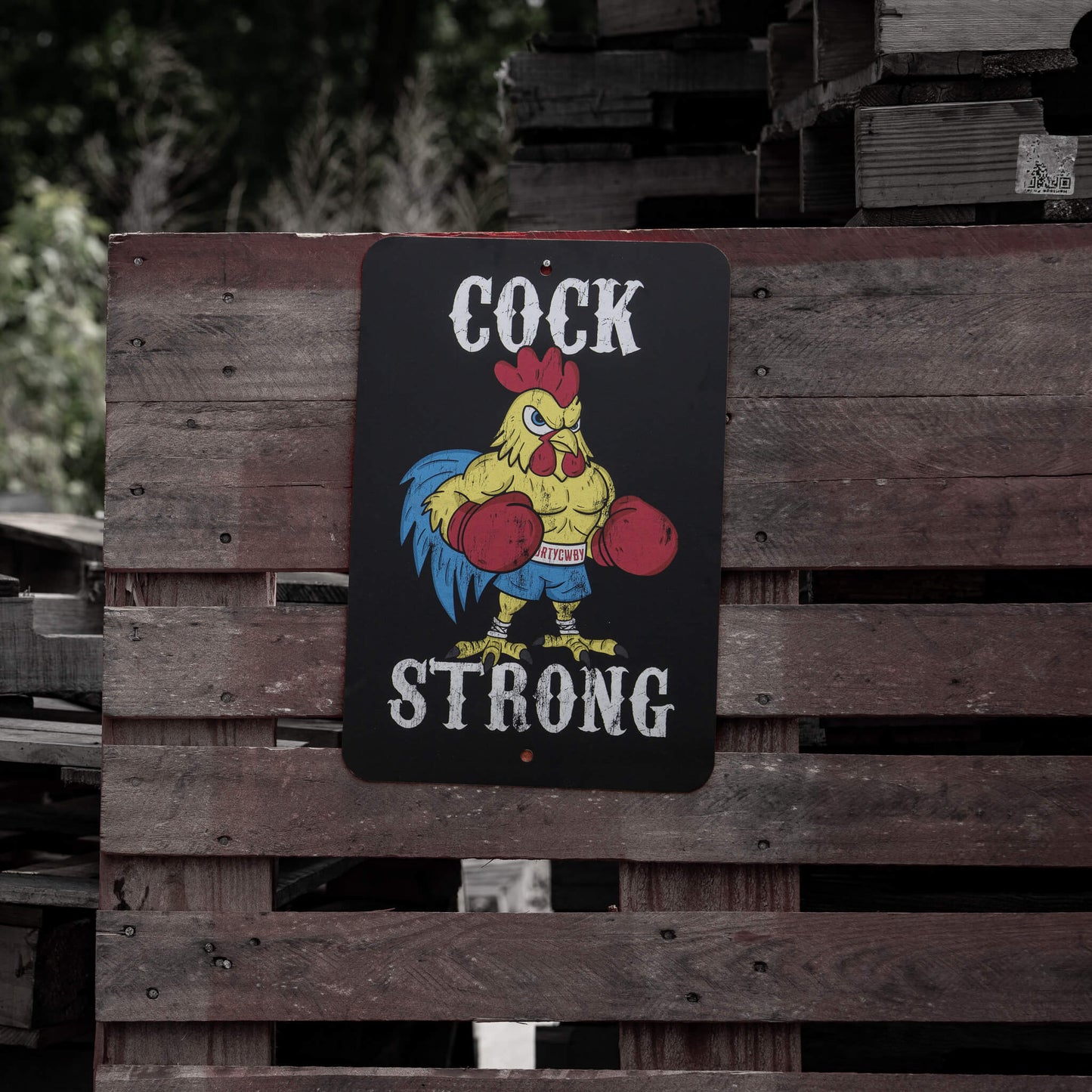 Cock Strong metal sign by Combat Iron Apparel displayed against wooden pallets, showcasing a tough-looking rooster.