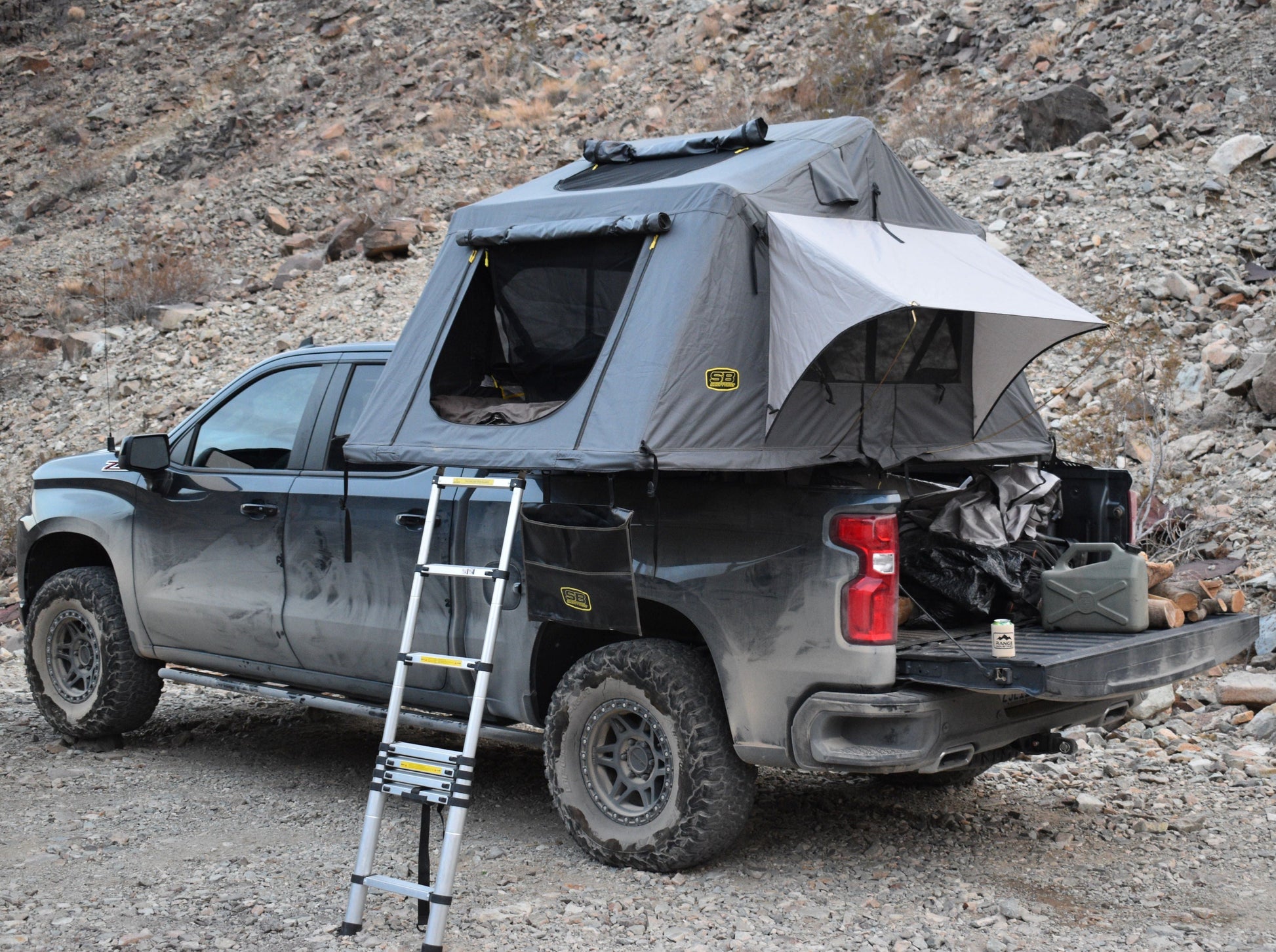 Range Industries 2019+ Silverado/Sierra 1500 Low Profile Tent Rack displayed on a truck with a rooftop tent setup, ladder leaning against the truck, set in a rocky outdoor environment.