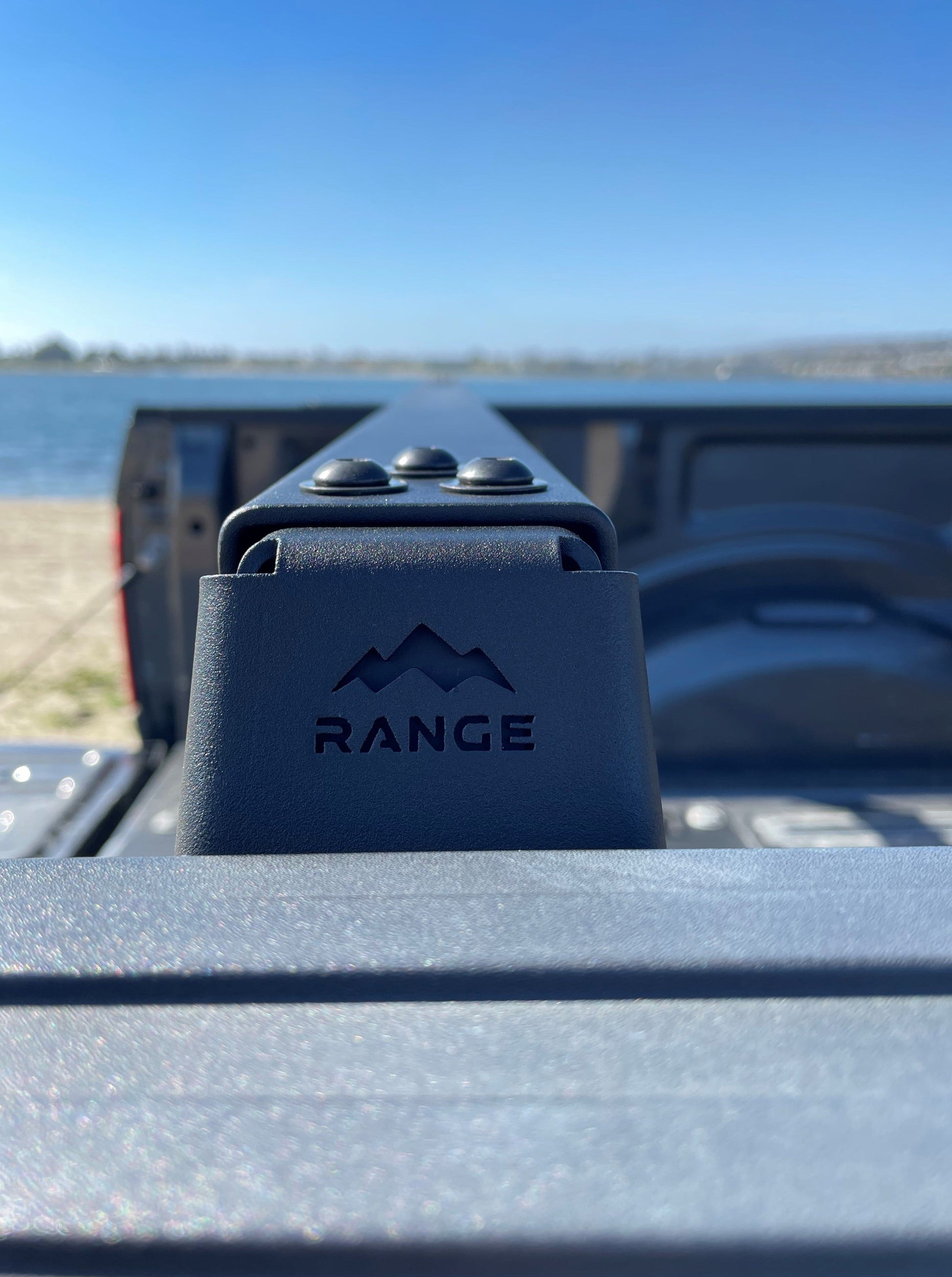 Close-up of the Range Industries logo on the mount of the 2015+ F-150 Low Profile Rooftop Tent Rack Kit, set against a backdrop of water and sky.