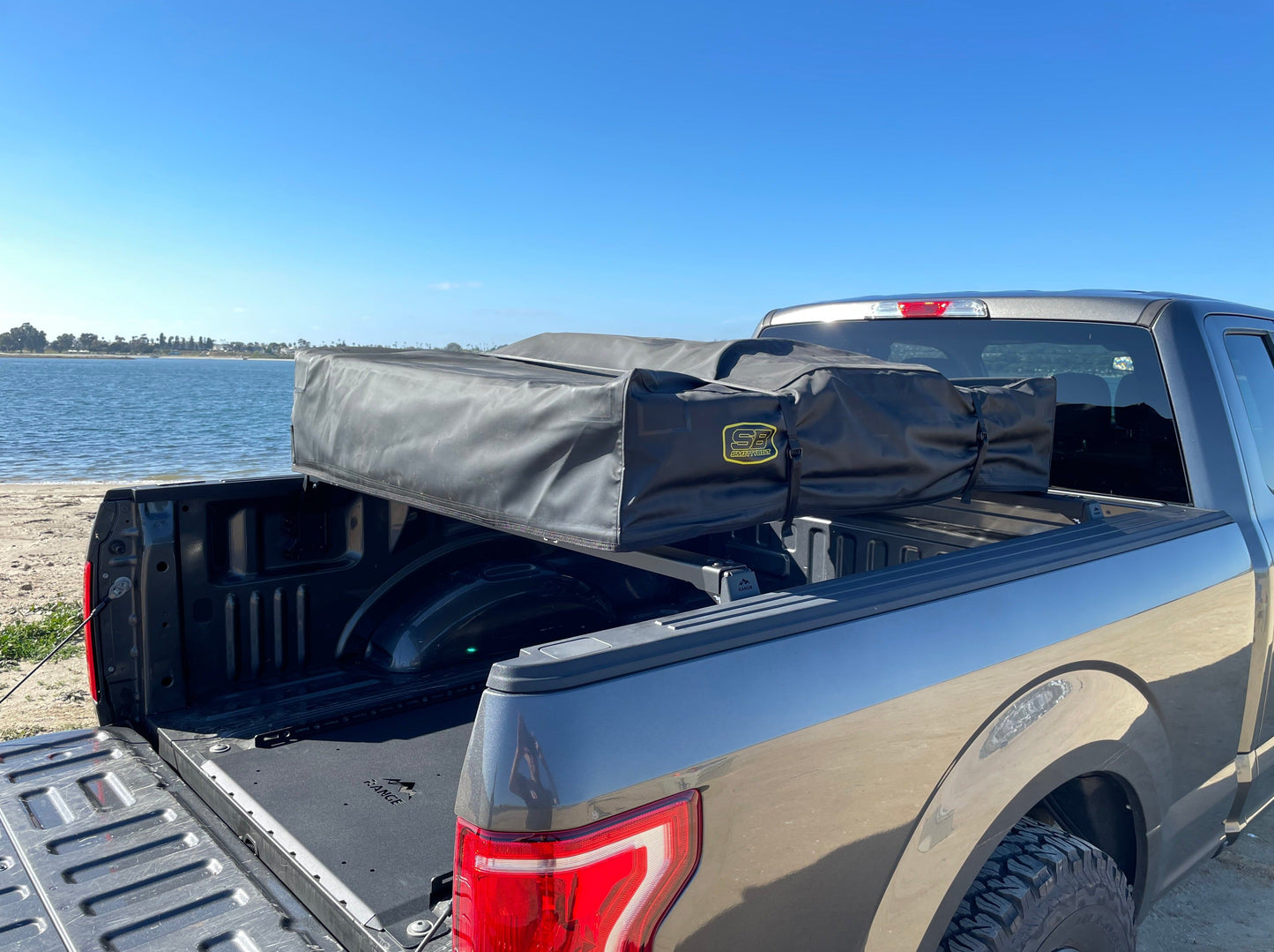 2015+ F-150 Low Profile Rooftop Tent Rack Kit by Range Industries, showcasing a close-up of the rooftop tent installed in the truck bed of a gray F-150, with a scenic beach view behind.