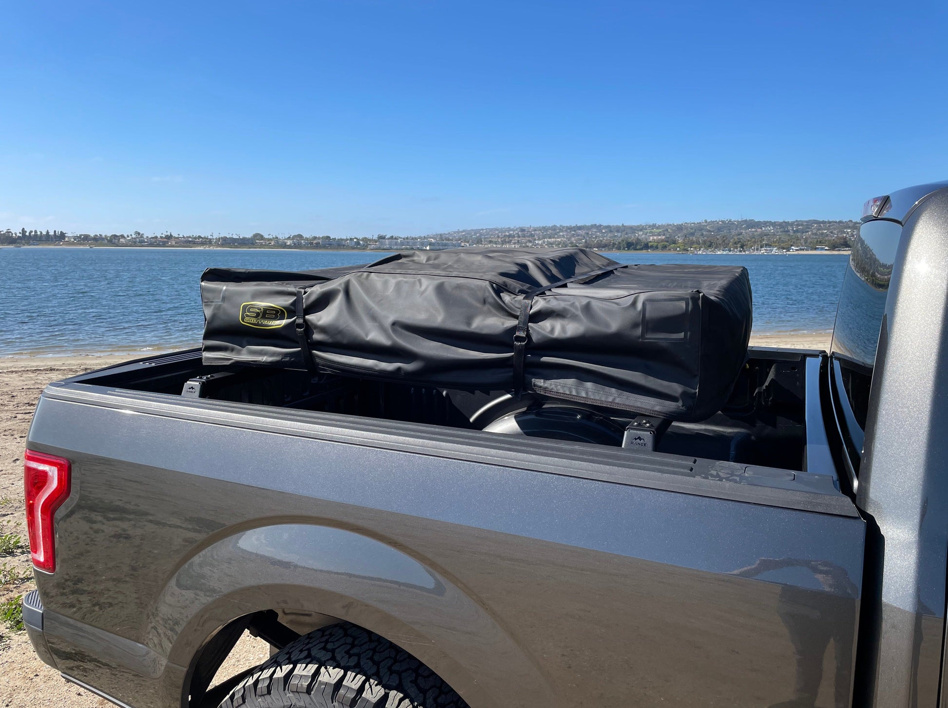 2015+ F-150 Low Profile Rooftop Tent Rack Kit by Range Industries, capturing the truck bed of a gray F-150 with a rooftop tent secured and a scenic waterfront in the background.