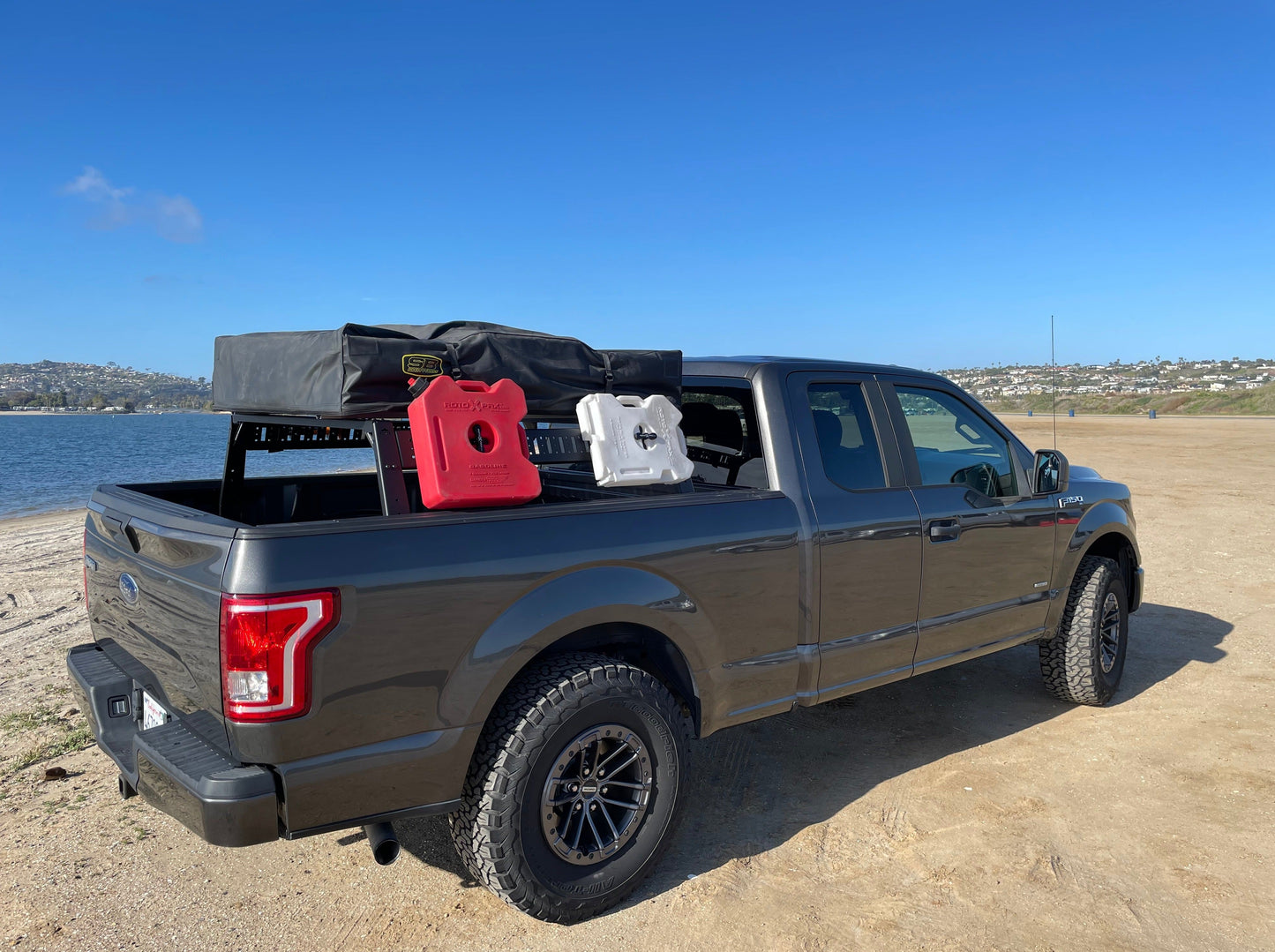 View of the Range Industries 2015+ F-150 Mid Height Rooftop Tent Rack Kit with accessories on a Ford F-150 parked on the beach, highlighting its functionality for outdoor adventures.