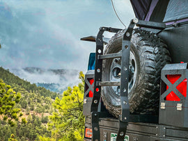 Dirtbox Overland Flatbed Ladder Upgrade showcasing a secure spare tire mount designed for tires up to 37 inches, surrounded by scenic mountain views.