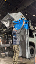 Load image into Gallery viewer, A user demonstrating the Ultimate Camp Shower Set by Dirtbox Overland, showcasing the hard case shower and bathroom awning being set up. The awning features a blue and gray design, perfect for outdoor adventures.