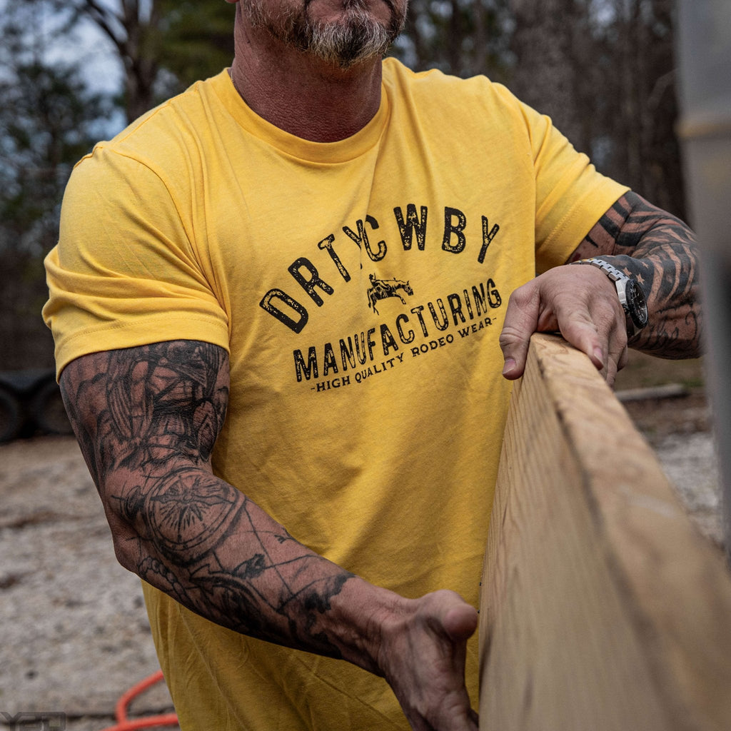 Muscular man wearing a bright yellow t-shirt that displays 'DRTYCWBY MANUFACTURING - HIGH QUALITY RODEO WEAR', representing the Original Dirty Cowboy MFG by Combat Iron Apparel.