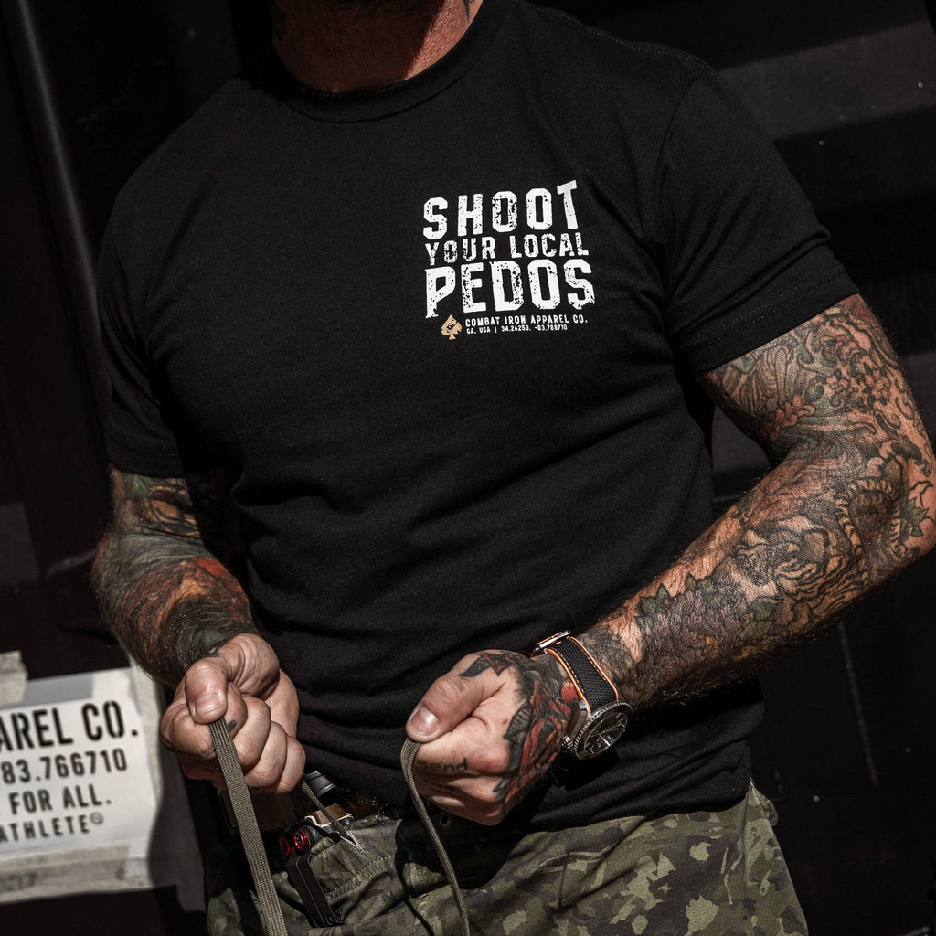 A casual portrait of a muscular man stretching in a black t-shirt with 'Shoot Your Local Pedos' on the back, from Combat Iron Apparel.
