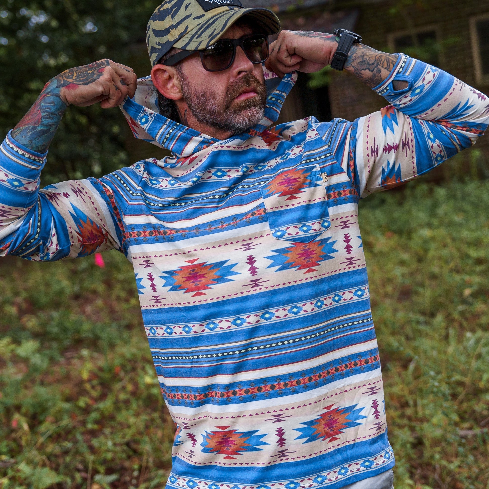A man in a tribal patterned Men's Outdoors Long Sleeve Performance Hoodie by Combat Iron Apparel, adjusting the hood outdoors.