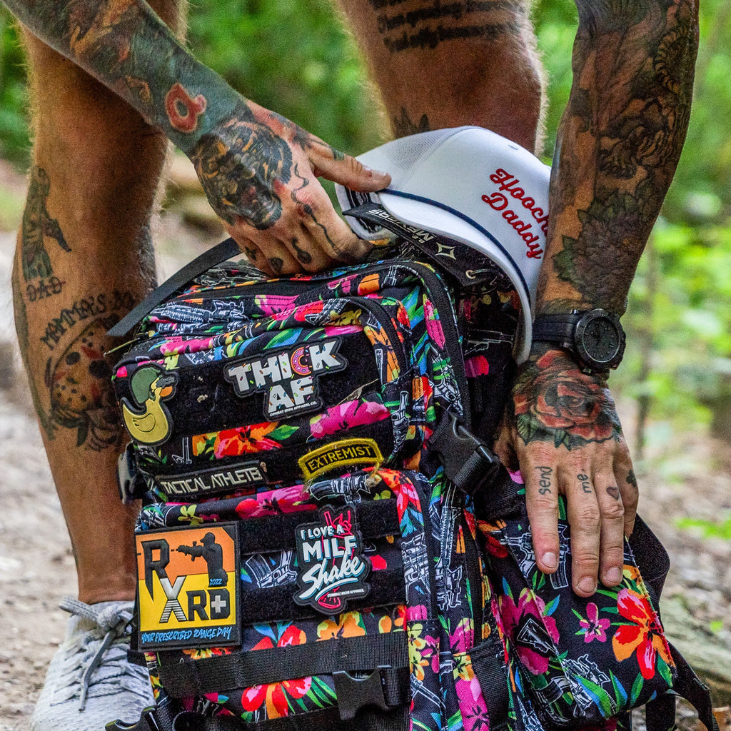 Close-up of the Combat Iron Apparel 25L Sack™ Original All-Day Molle Backpack with Pouch, featuring colorful patches on a floral background.
