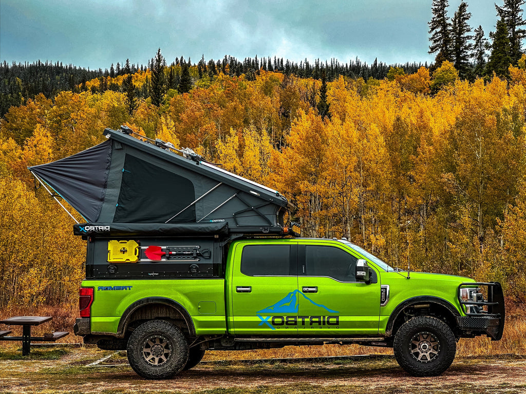 Vibrant green truck equipped with a Genesis 4‑Season Insulated Overland Camper Vehicle by Dirtbox Overland, featuring a rooftop tent, parked against a backdrop of colorful autumn trees. Perfect for rugged off-grid adventures.