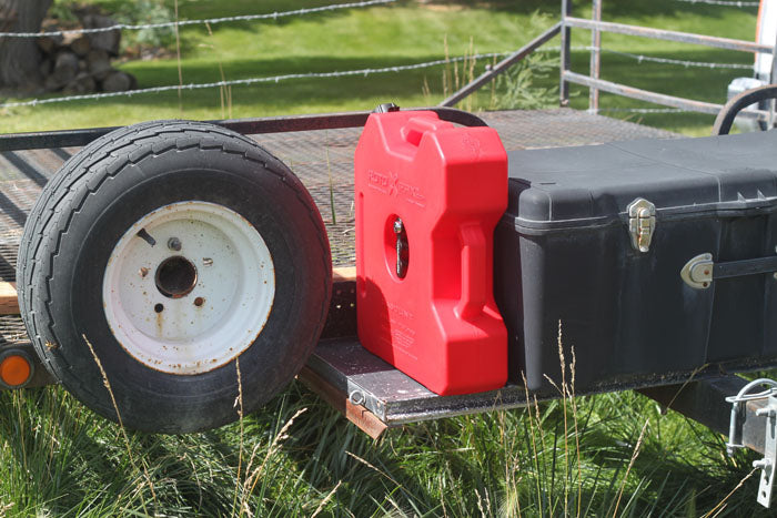 A red RotopaX 3 Gallon Extension fuel container positioned on a trailer next to a black storage box and a wheel, showcasing its durable design and practical use for stacking fuel packs.