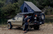 Load image into Gallery viewer, Dirtbox Overland Flatbed System Full Box installed on a truck by a river, showcasing its rugged camping features and interior lighting.