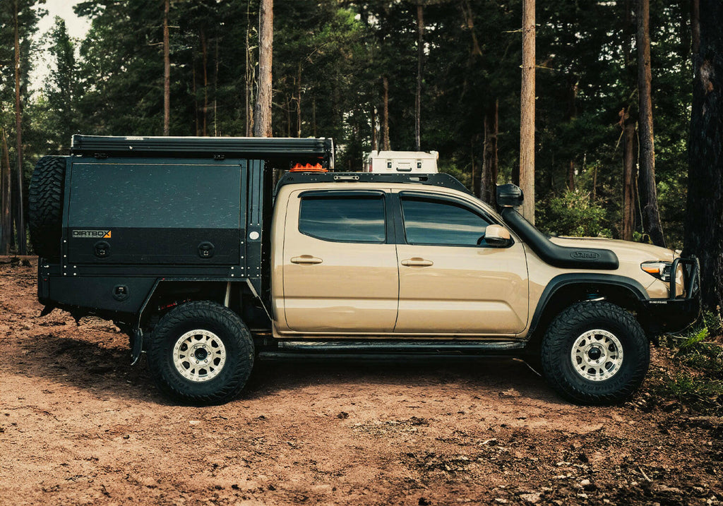Side view of a Dirtbox Overland Flatbed System Full Box mounted on a beige truck in a forested area, illustrating its versatile design and rugged appearance.