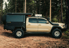 Load image into Gallery viewer, Side view of a Dirtbox Overland Flatbed System Full Box mounted on a beige truck in a forested area, illustrating its versatile design and rugged appearance.
