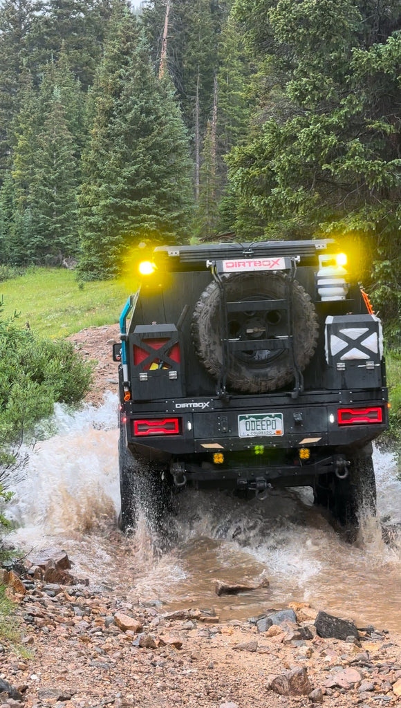 Off-roading adventure featuring a Dirtbox Overland Flatbed System Full Box truck splashing through water in a forest setting. Perfect for outdoor enthusiasts looking for rugged versatility.